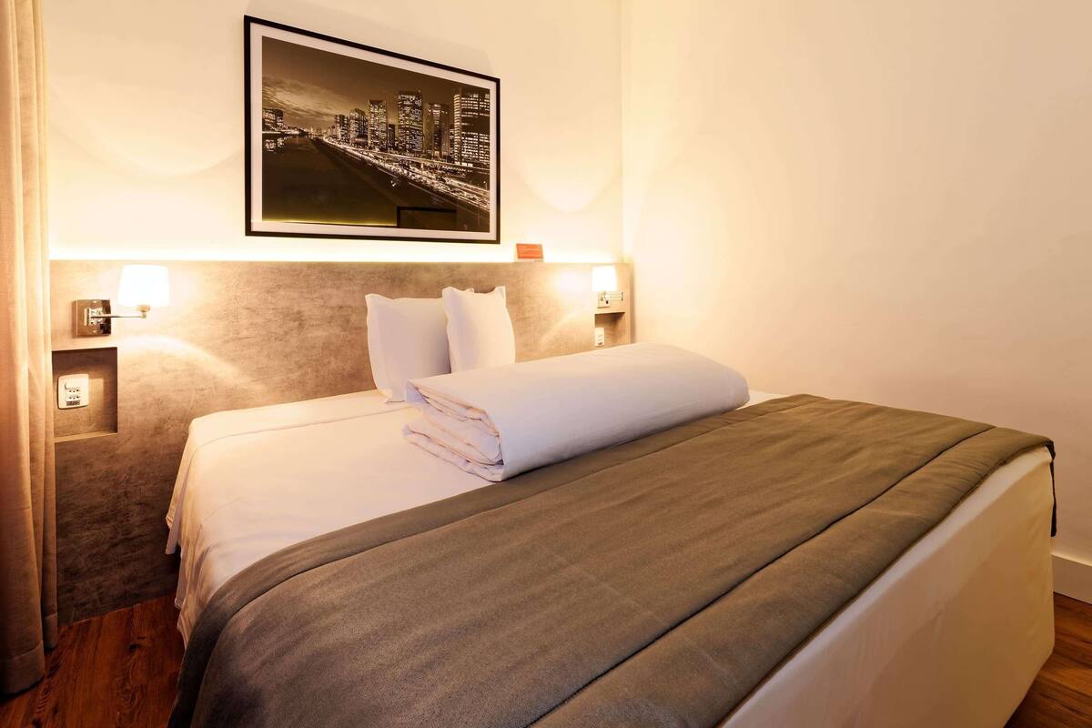A hotel room with a neatly made bed, a cityscape picture on the wall, and two bedside lamps.