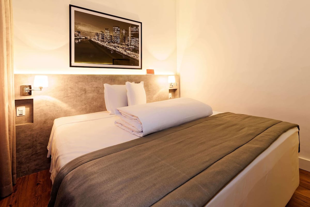 Une chambre d’hôtel avec un lit soigneusement fait, un tableau représentant un paysage urbain accroché au mur et deux lampes de chevet.