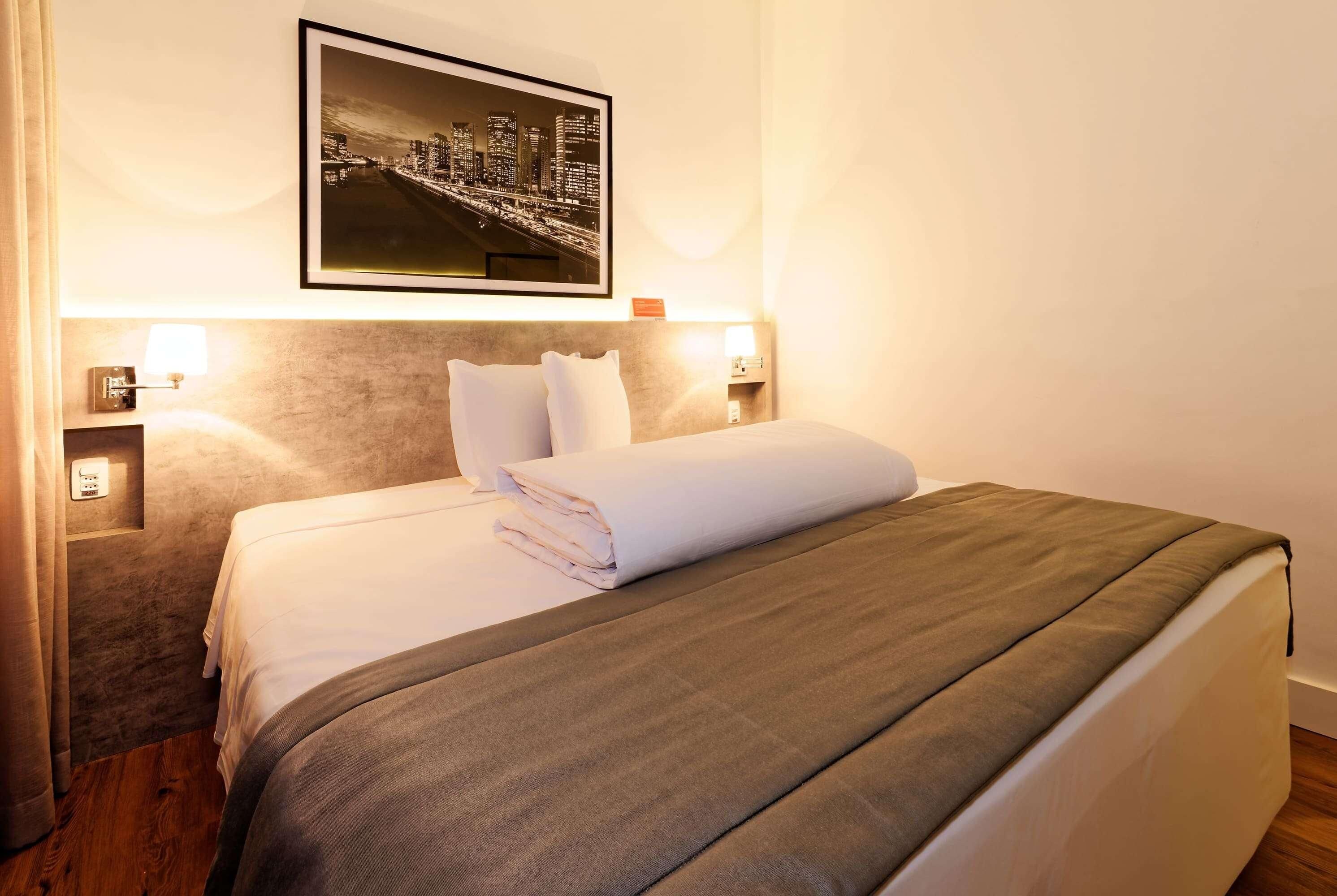 Uma cama bem arrumada com um cobertor cinzento, uma fotografia emoldurada da paisagem urbana acima dela e abajures de cada lado.