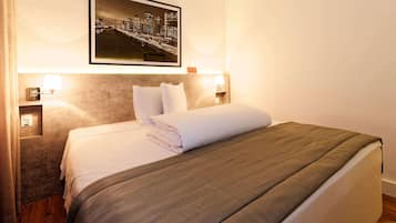 Uma cama bem arrumada com um cobertor cinzento, uma fotografia emoldurada da paisagem urbana acima dela e abajures de cada lado.