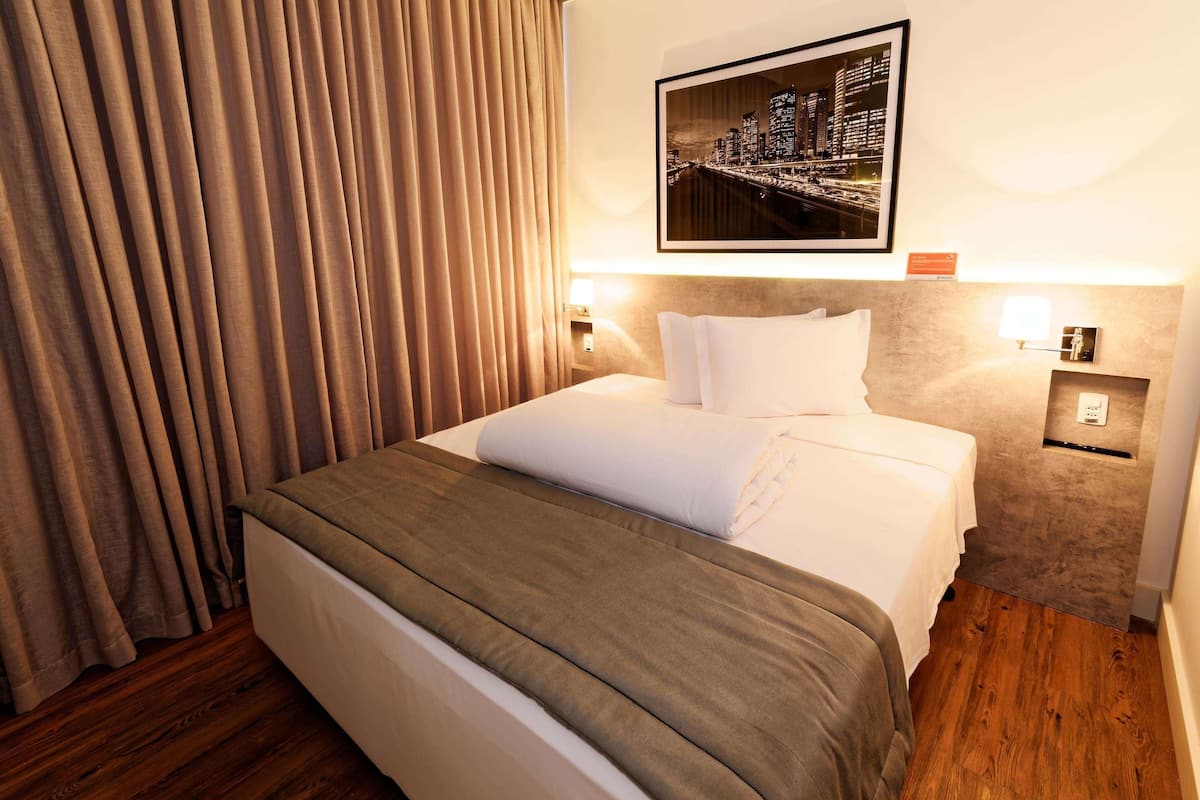 A hotel room with a neatly made bed, a cityscape picture on the wall, and a wooden floor.