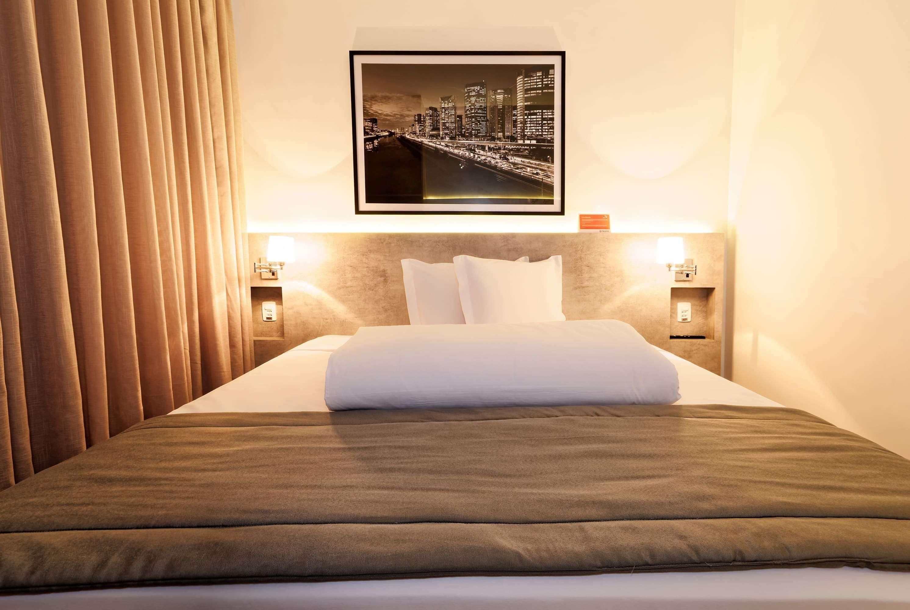 Quarto de hotel com uma cama bem arrumada, um quadro com vista urbana na parede e duas luminárias de cabeceira.