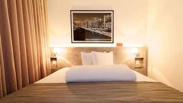 Quarto de hotel com uma cama bem arrumada, um quadro com vista urbana na parede e duas luminárias de cabeceira.