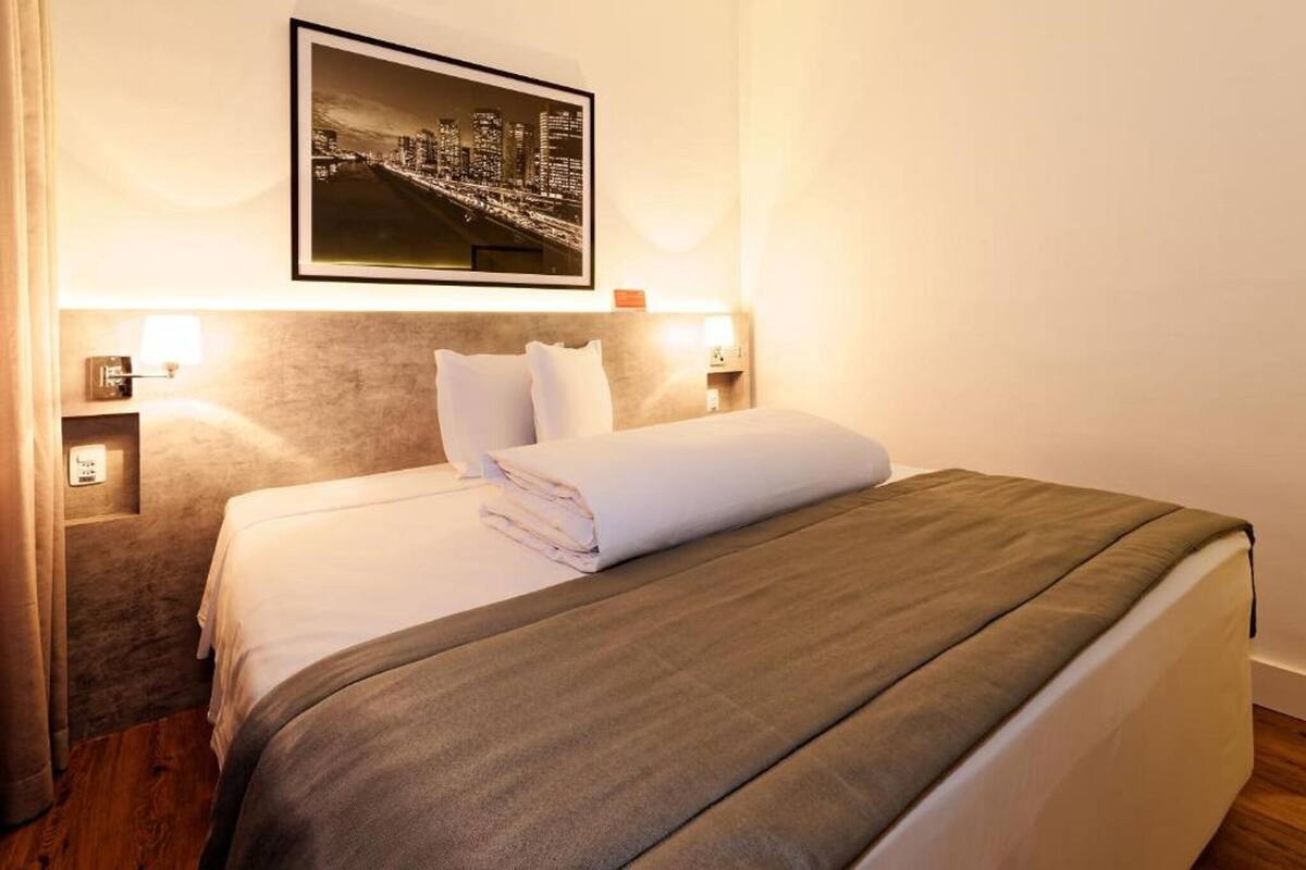 A hotel room with a neatly made bed, a framed cityscape picture on the wall, and two bedside lamps.