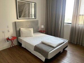 Quarto de hotel com uma cama grande, uma mesinha de cabeceira com telefone, uma mesa vermelha e uma janela com cortinas.