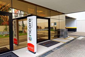 Entrada de hotel com fachada de vidro, placa indicando hotel Encore e um carrinho estacionado na frente.