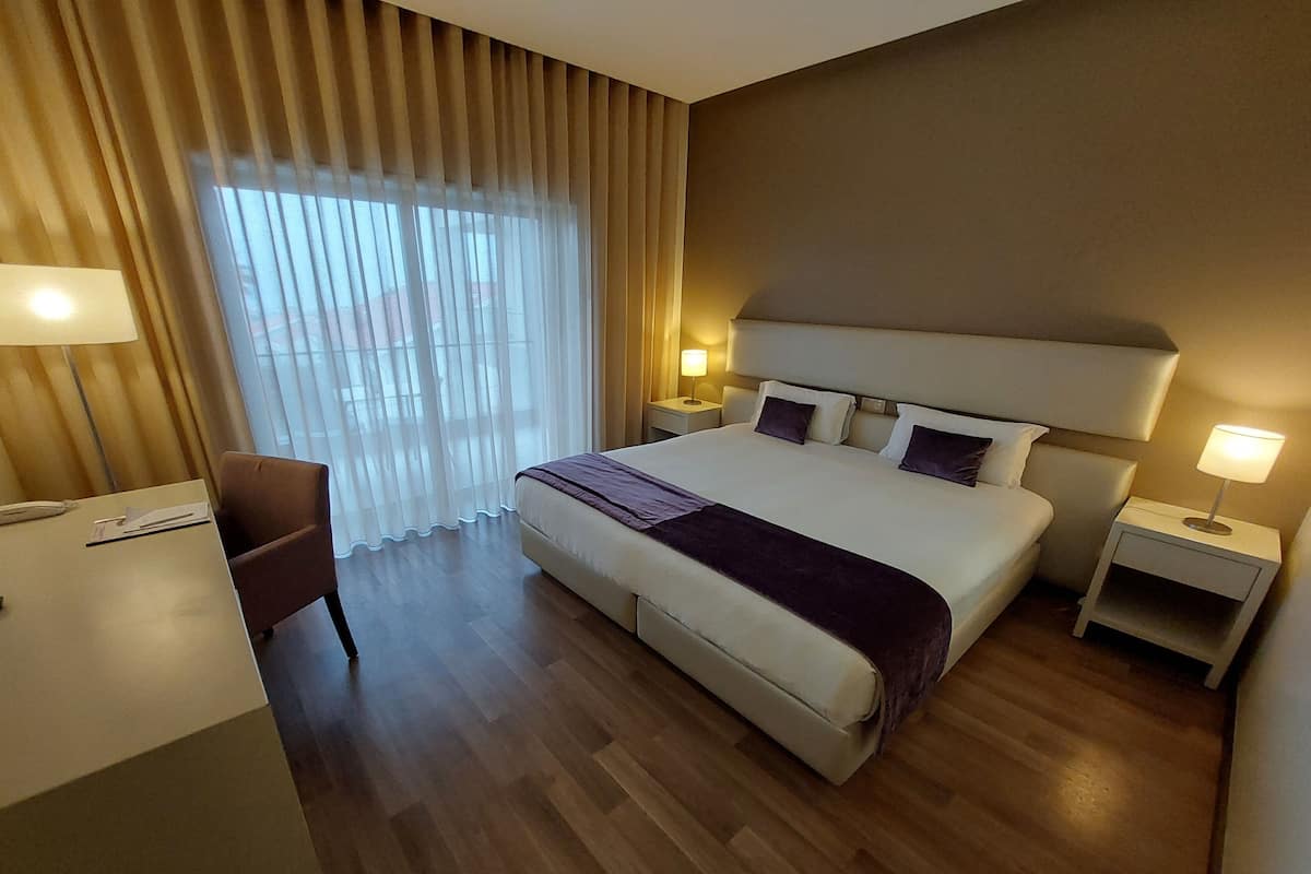 Habitación de hotel con una cama grande, dos mesitas de noche con lámparas, un escritorio con una silla y una ventana con cortinas translúcidas.