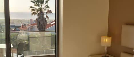 A hotel room with a bed, a nightstand with a lamp, a chair, and a balcony with a view of the sea and palm trees.