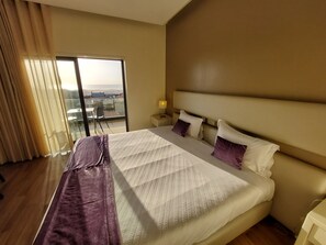 A hotel room with a large bed, a view through a sliding door, and a nightstand with a lamp.