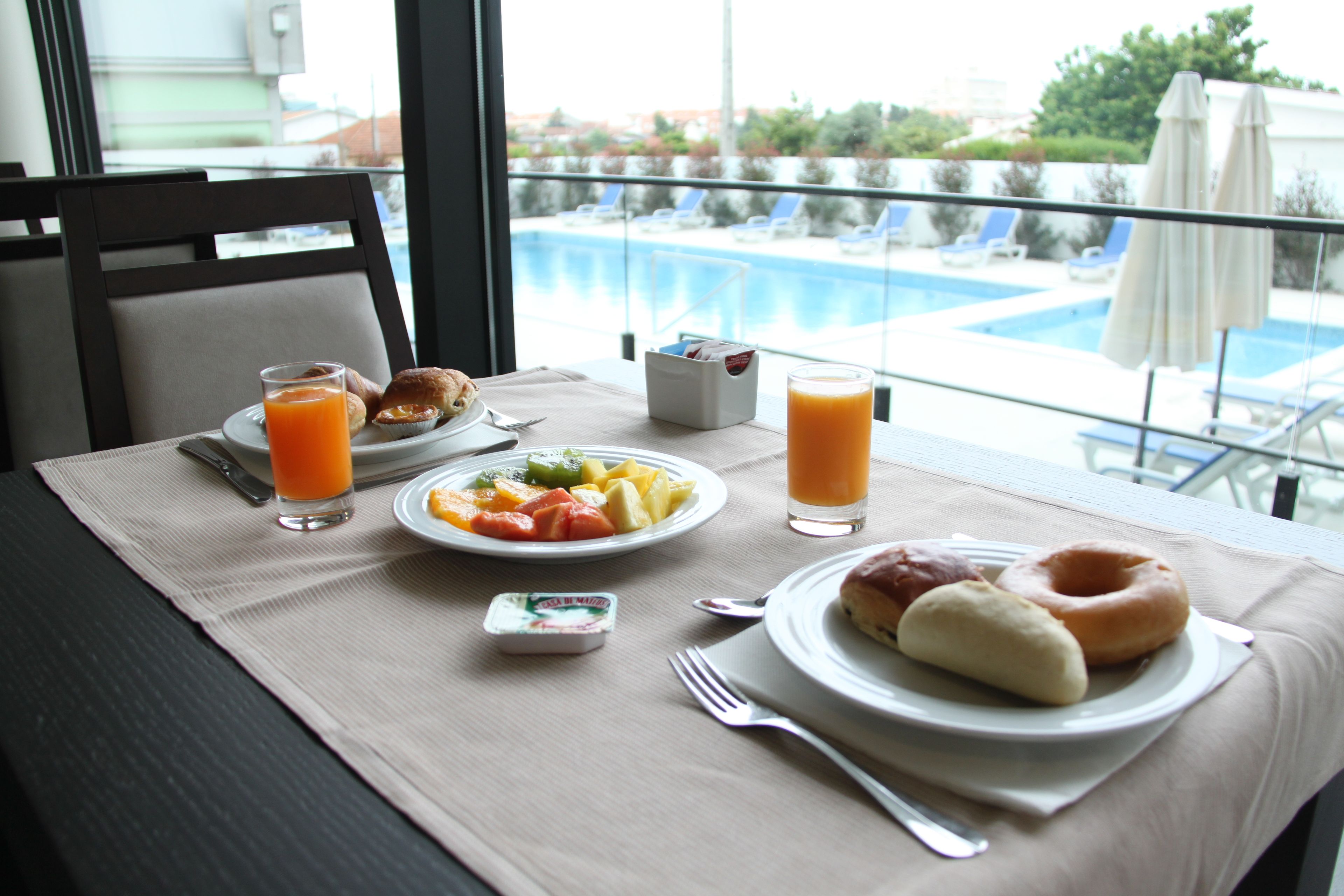 Et morgenbord med tallerkener med mad, en frugtskål og glas appelsinjuice, med udsigt til en swimmingpool.