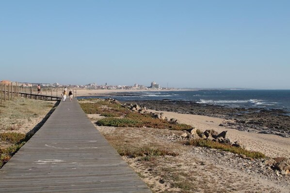 A wooden boardwalk leading to a beach with rocky terrain and a distant cityscape.
