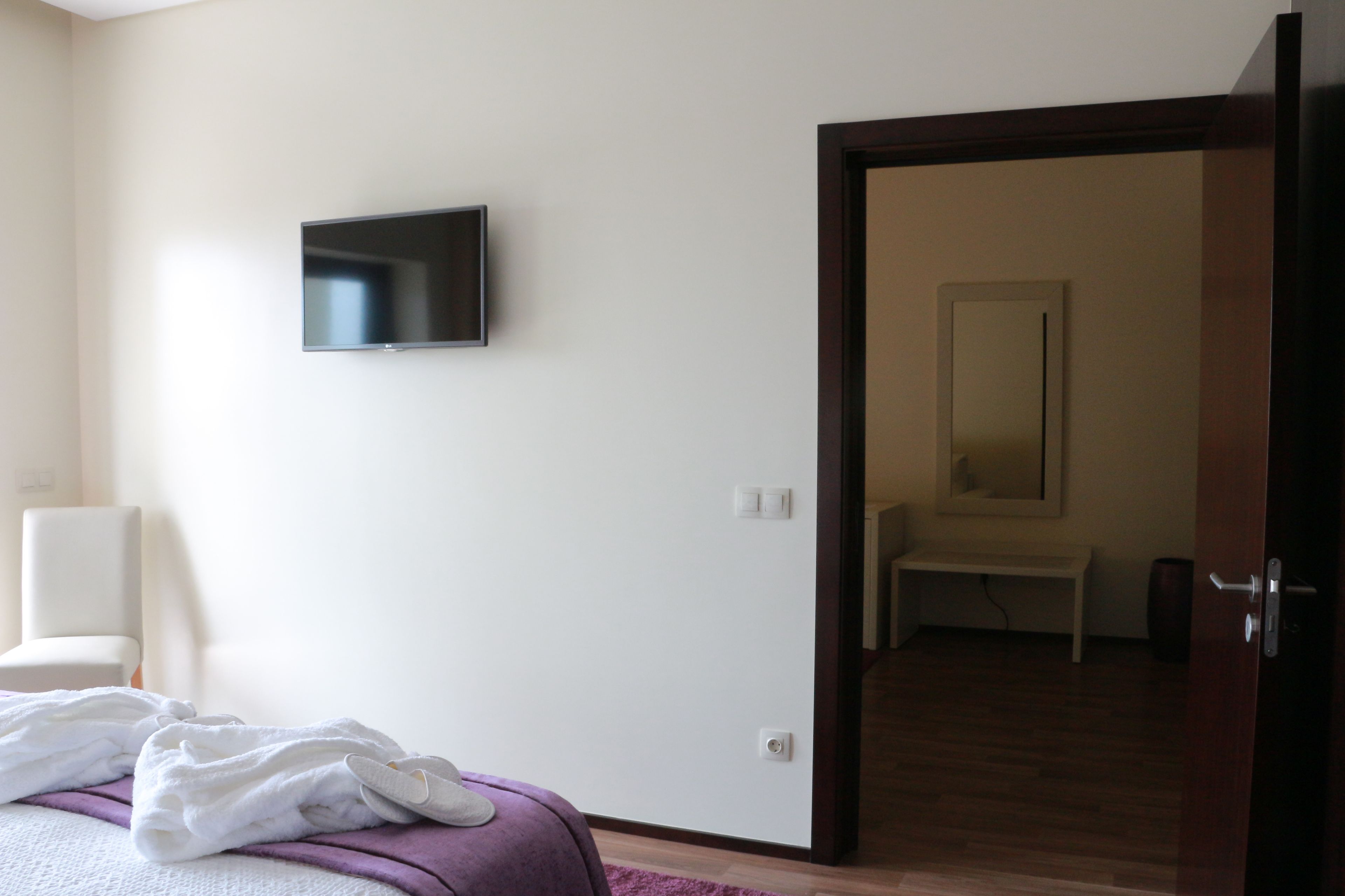 Quarto com cama, televisão fixada na parede e porta de acesso a um banheiro.