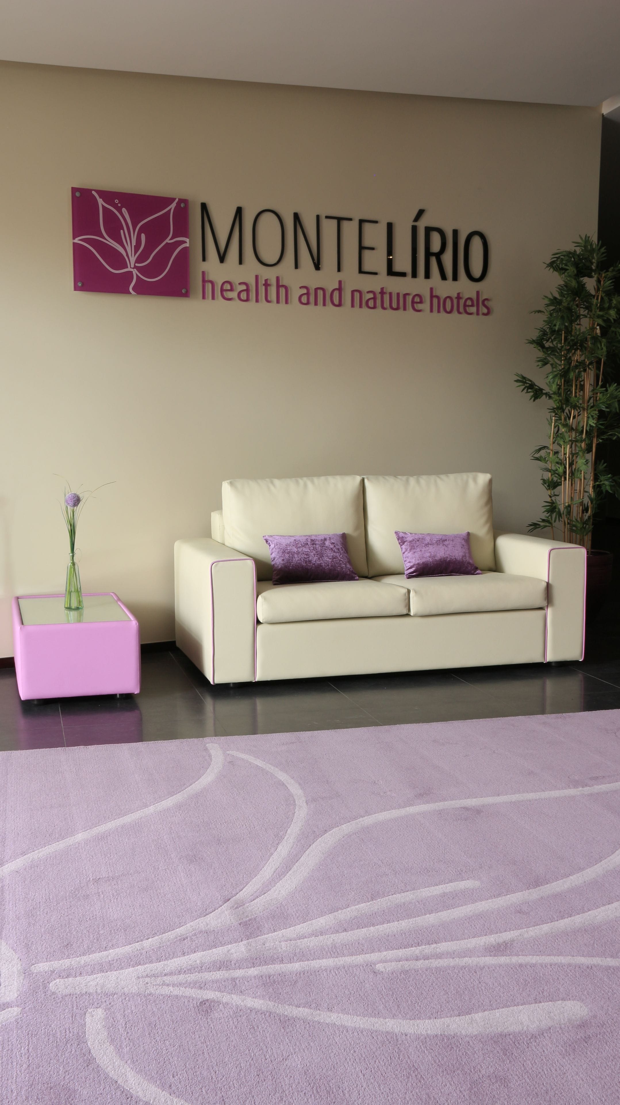 A modern hotel room with a beige sofa, purple cushions, a pink side table, and a purple rug.