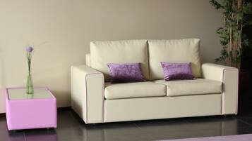 A modern hotel room with a beige sofa, purple cushions, a pink side table, and a purple rug.