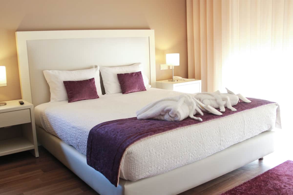 Una cama bien hecha con sábanas blancas y detalles morados, dos mesitas de noche con lámparas y un suelo de madera.