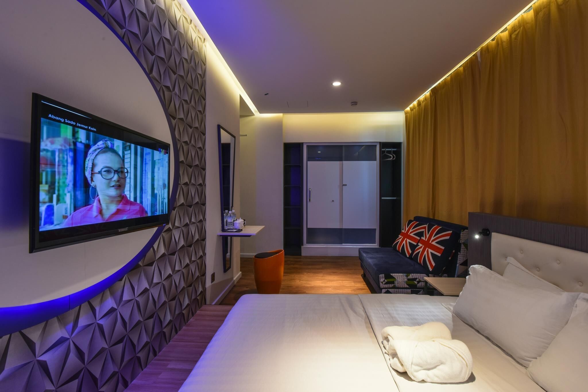 A modern hotel room with a large TV mounted on the wall, a bed with white linens, a bedside table, and a wardrobe.