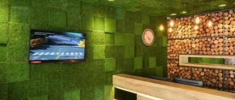 Un espace d’accueil avec un mur texturé de couleur verte, un comptoir d’accueil en bois, une horloge et un téléviseur fixé au mur.