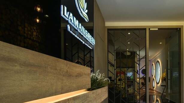 Zona de recepción con mostrador de madera, un letrero que dice "ILAMANGREEN" y un interior moderno con suelo de madera en espiga y puertas de vidrio que dan a un vestíbulo.