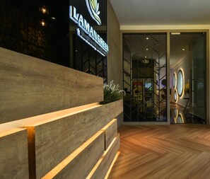 Un espace d’accueil doté d’un comptoir en bois, d’une enseigne indiquant « ILAMANGREEN », et d’un intérieur moderne avec un parquet en bois disposé en chevrons et des portes en verre donnant sur un hall d’entrée.