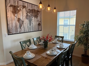 Un coin repas avec une table en bois dressée pour quatre personnes, entourée de chaises. Un grand tableau abstrait orne le mur, et il y a une fenêtre avec des stores.