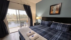 A hotel room with a bed, a nightstand, a lamp, a painting, and a view of a balcony with a table and chairs. - Oakwater, 3 bedrooms - The nearest resort from Disney! (Kissimmee)