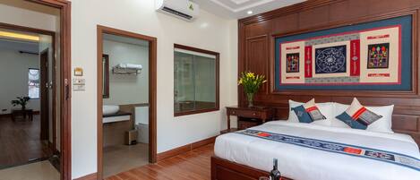 A hotel room with a large bed, wooden headboard, a wall-mounted air conditioner, a small table with a fruit bowl, and a magazine.