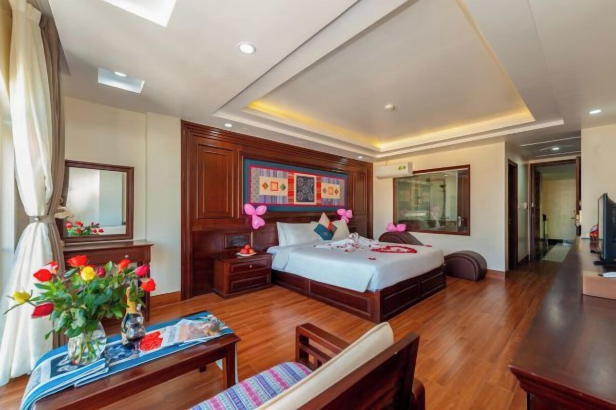 A hotel room with a large bed, a wooden nightstand, a desk with a chair, and a window with curtains.