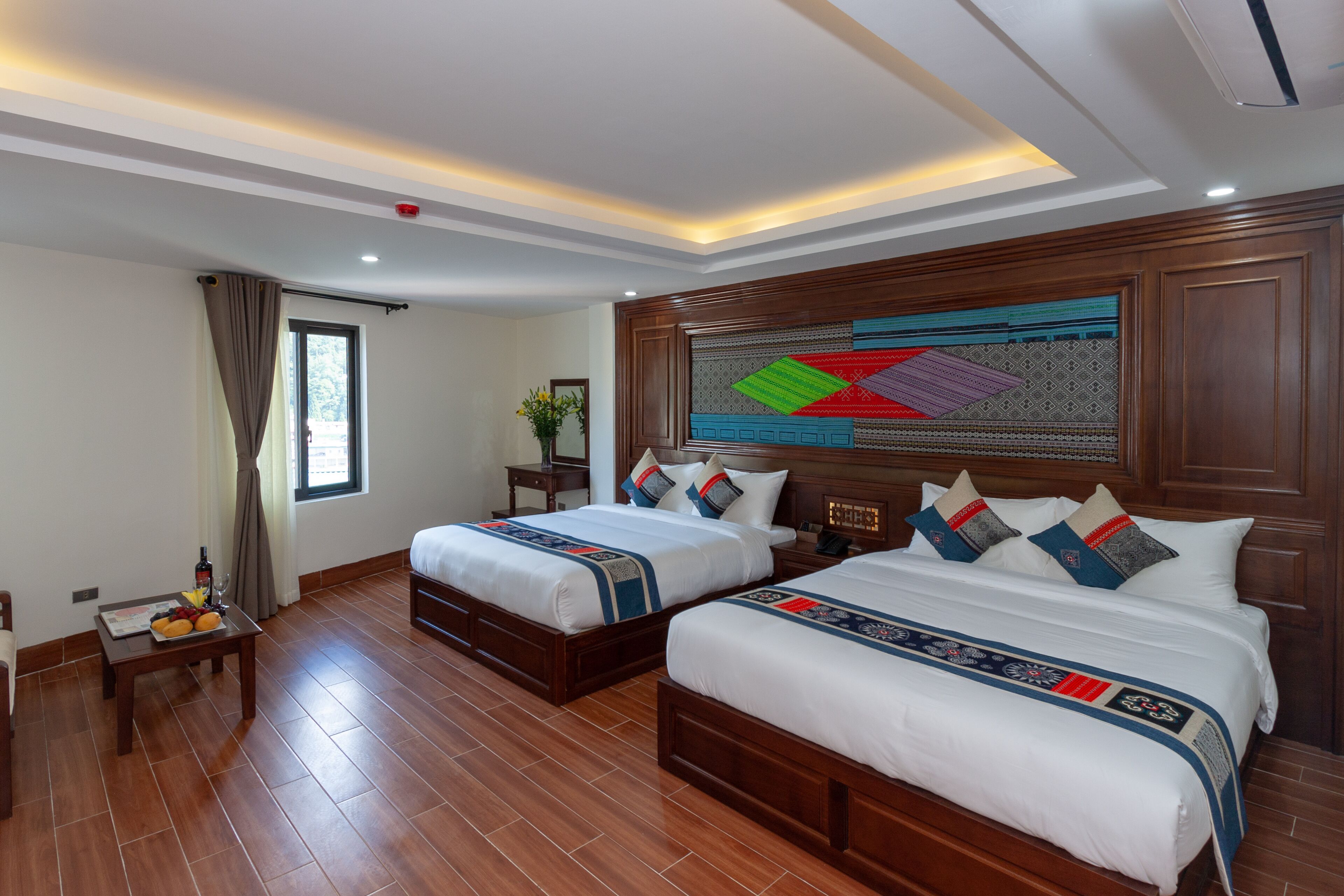 A hotel room with two beds, wooden headboard, a large window with curtains, a small table with fruit and a bottle, and a decorative wall hanging.