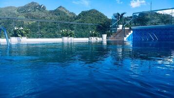 Piscina con agua azul cristalina, rodeada de macetas con plantas y vistas a colinas verdes.