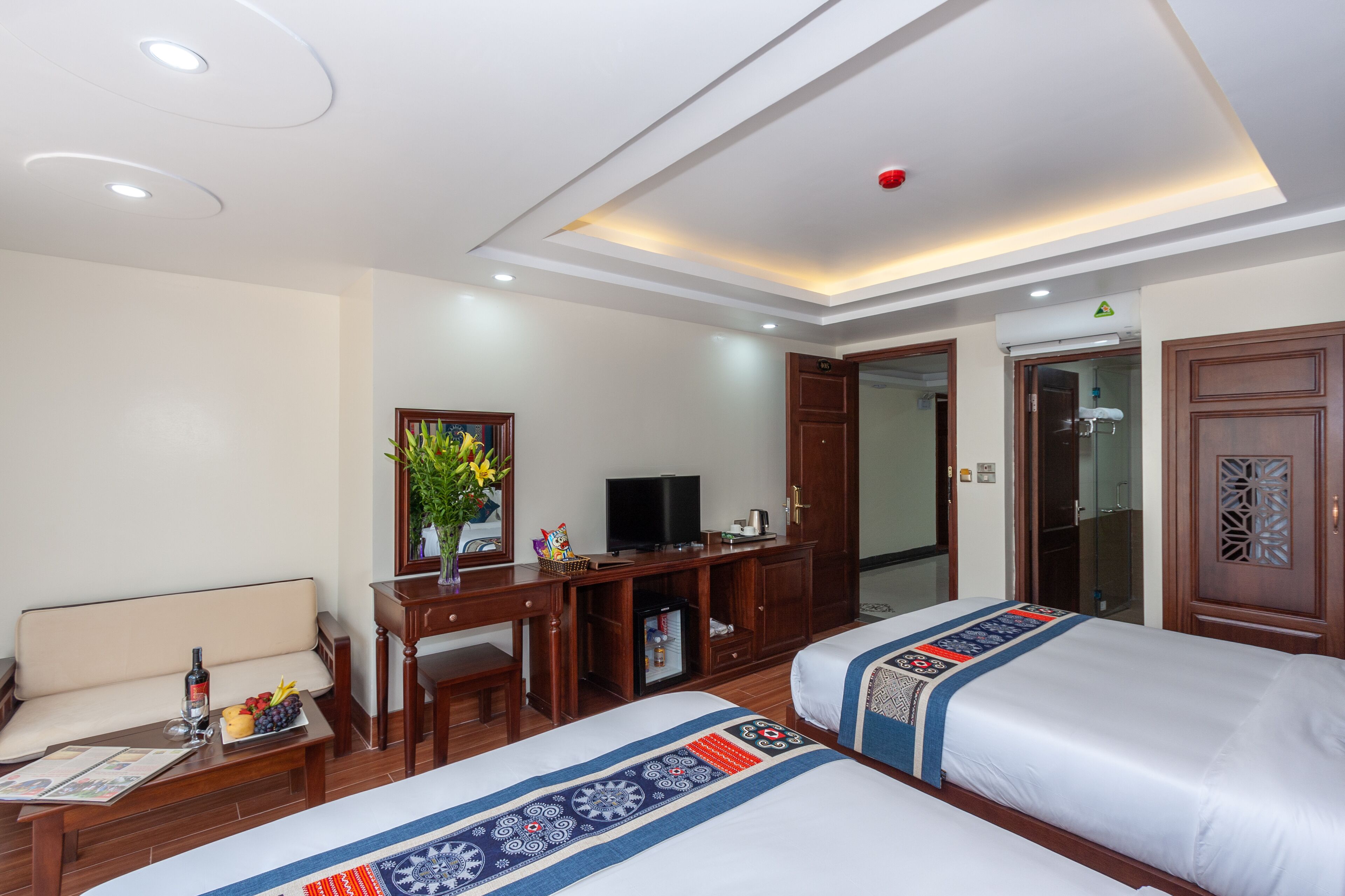 Habitación de hotel con dos camas, un escritorio con televisor, un sofá y una pequeña mesa con una botella y fruta.