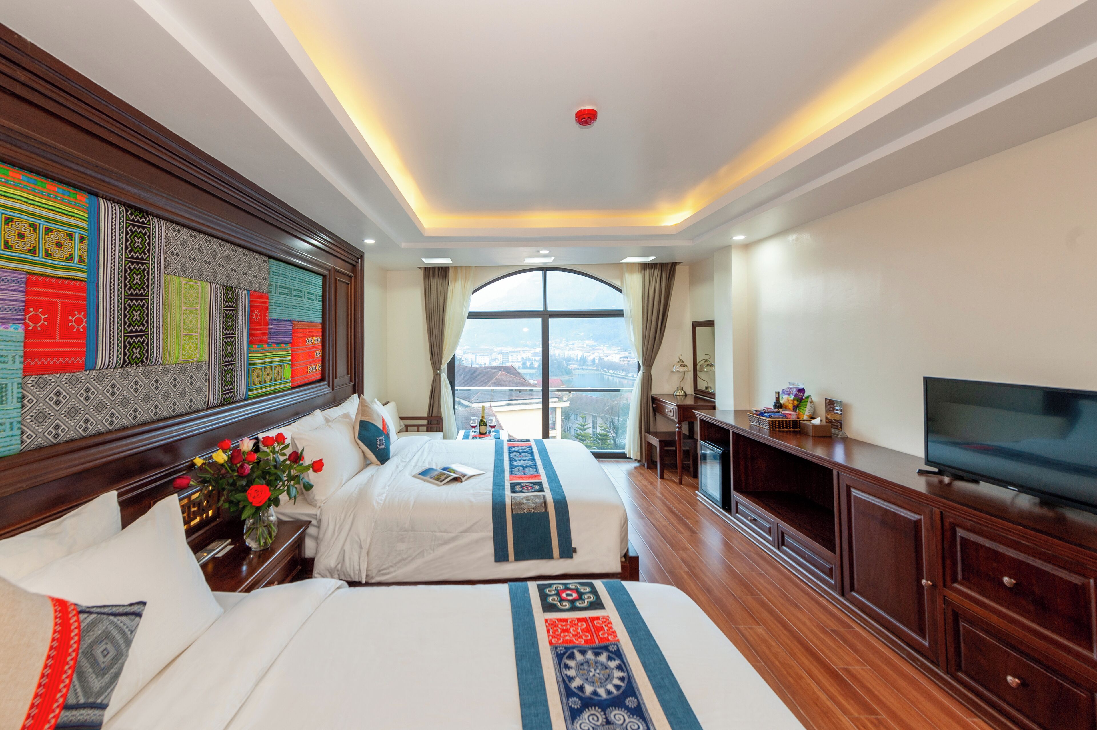 A hotel room with two beds, a TV, wooden furniture, a large window with a city view, and a decorative wall.