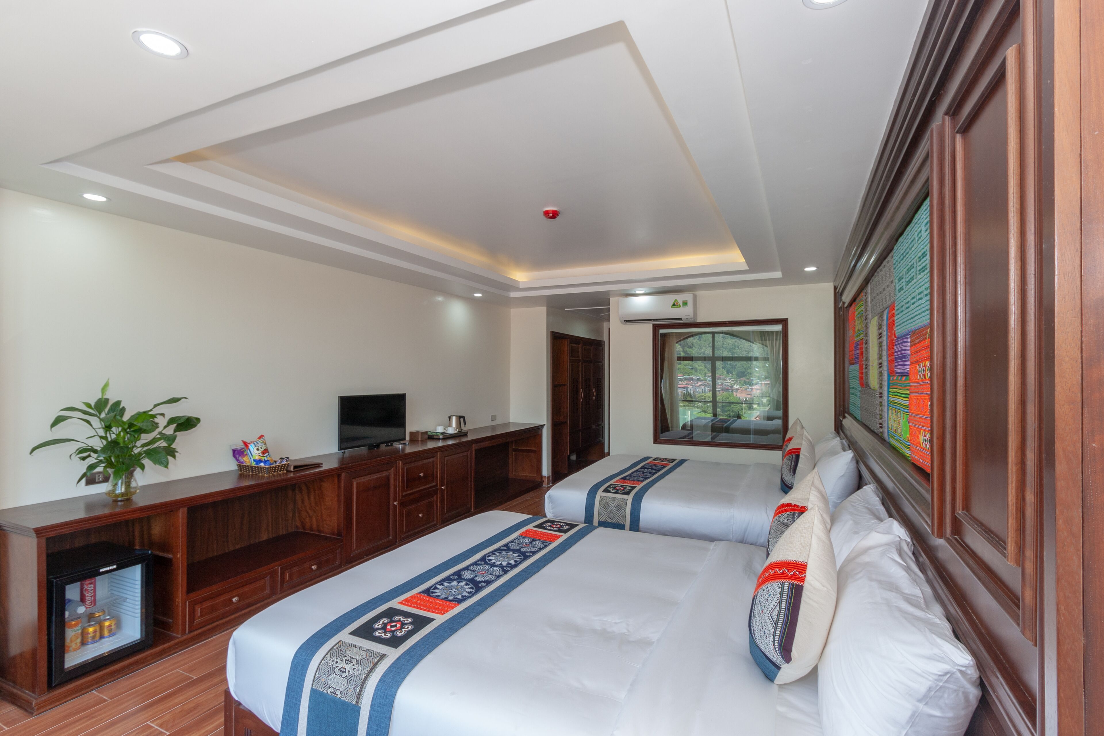 Habitación de hotel con dos camas, televisión, refrigerador pequeño y ventana con vistas.