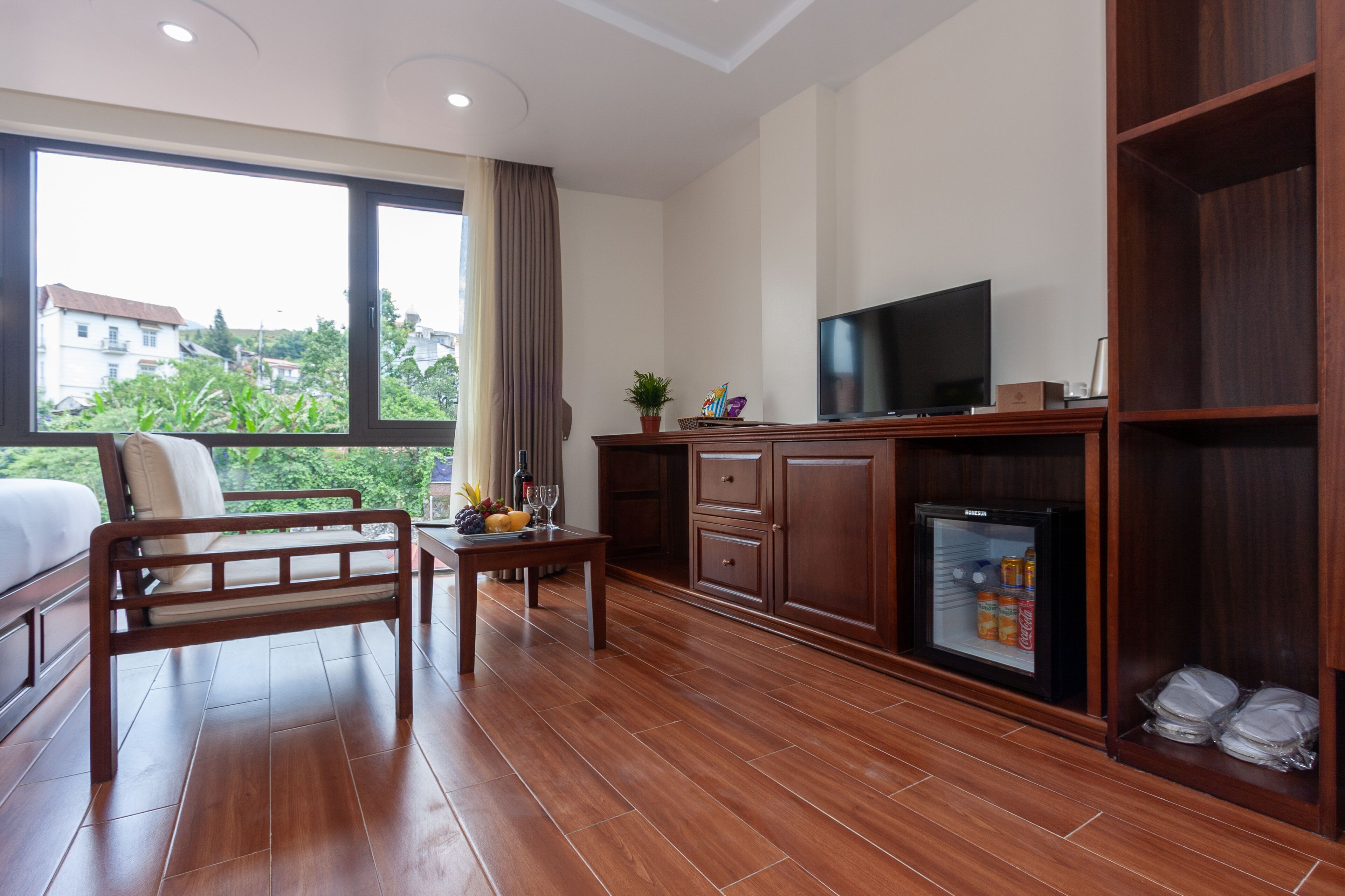 A hotel room with a bed, a wooden desk, a television, a mini-fridge, and a view of the outside.