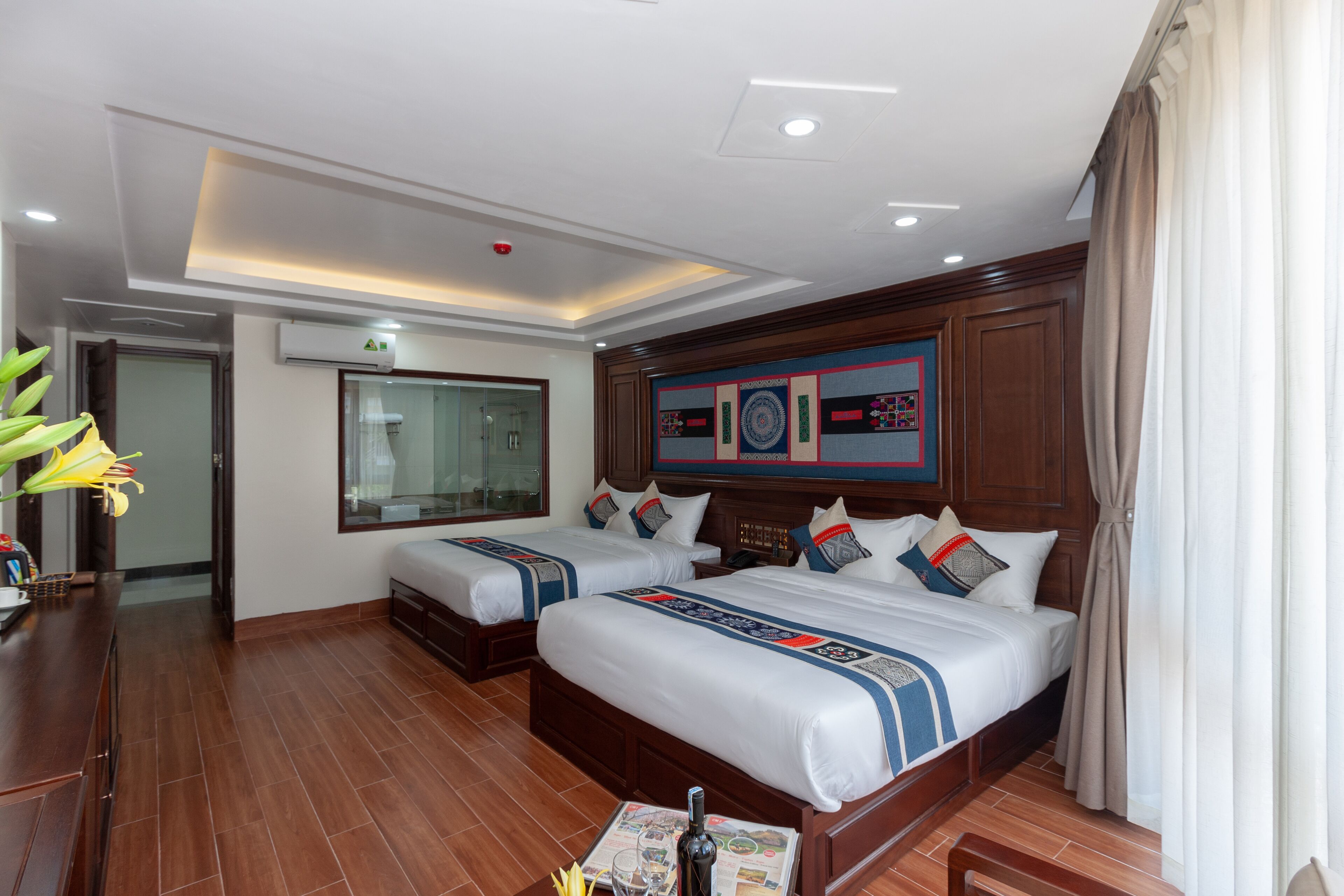 A hotel room with two beds, a wooden floor, a desk with a chair, a mirror, and a wall-mounted air conditioner.