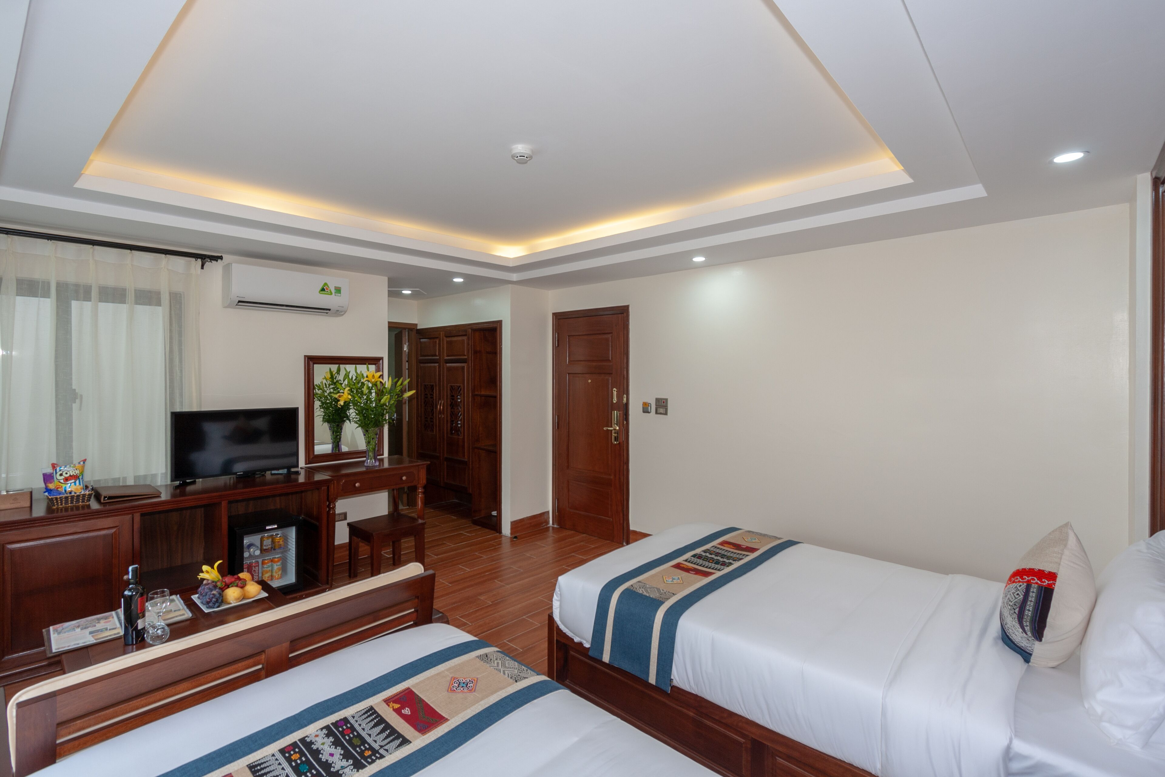Ein Hotelzimmer mit zwei Betten, einem Schreibtisch, einem Fernseher, einer Minibar und einem großen Fenster mit Vorhängen.
