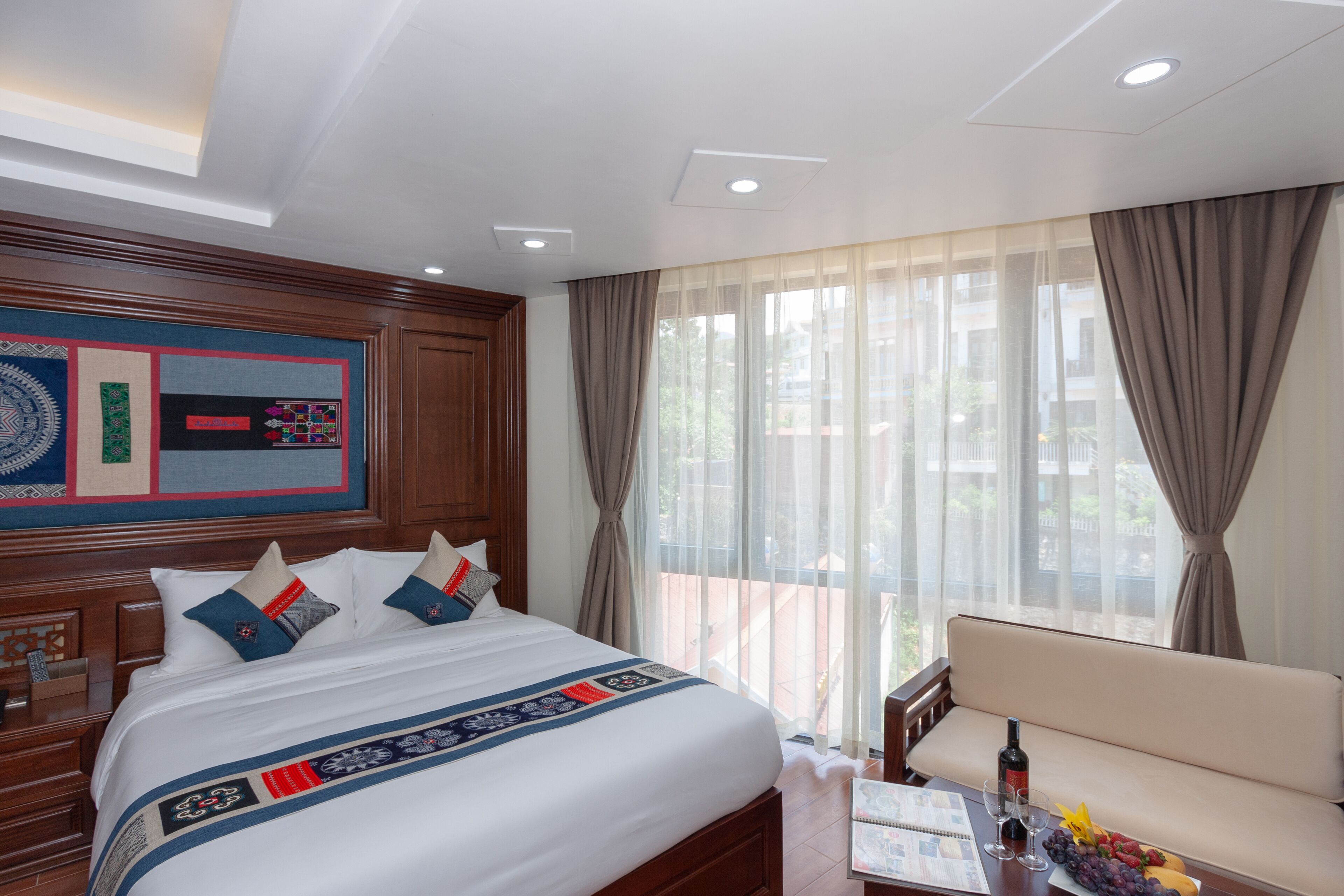 Habitación de hotel con una cama grande, cabecero de madera, ventana con cortinas, un sofá y una pequeña mesa con una botella y fruta.