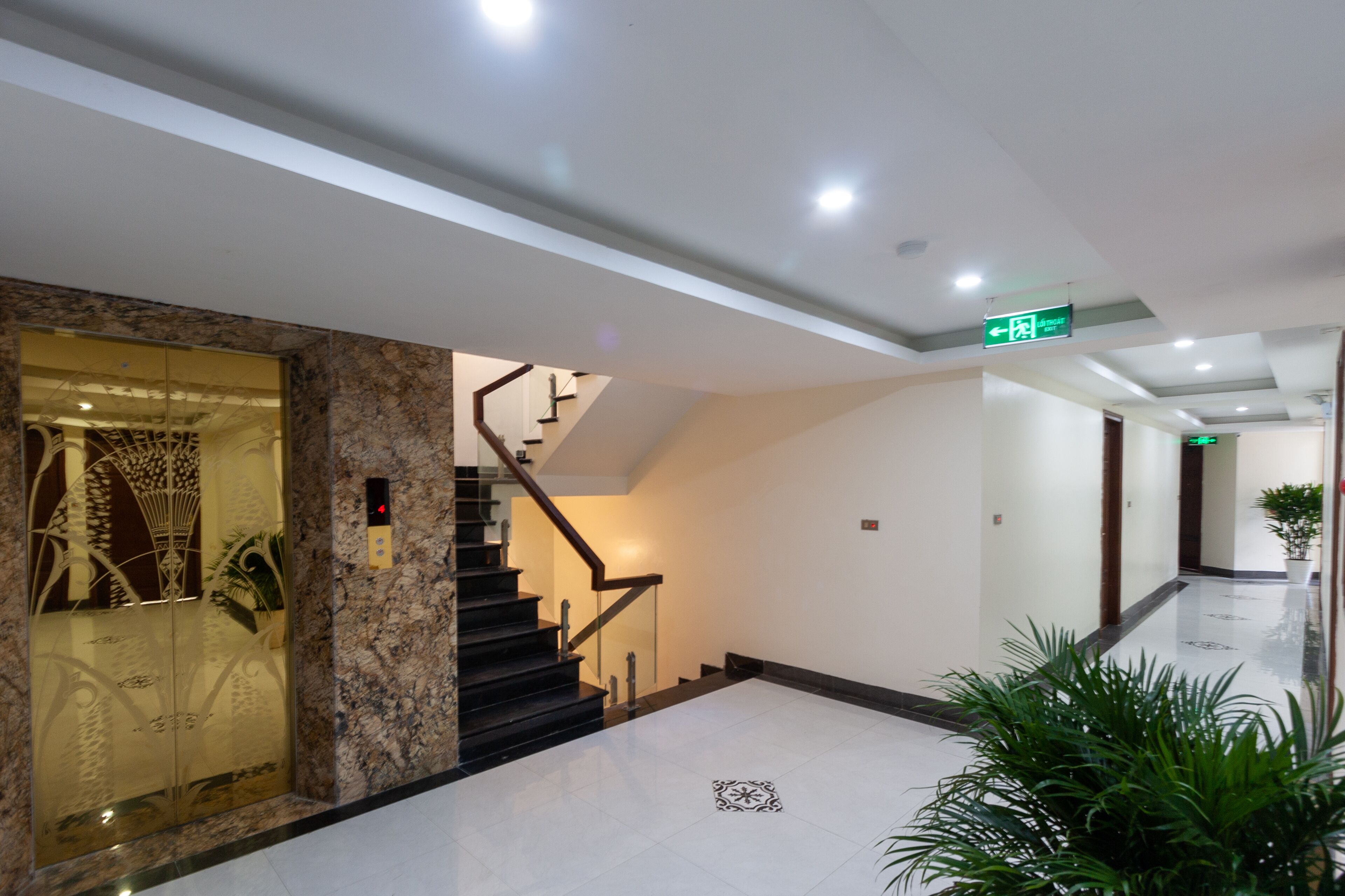 A modern hotel lobby with a marble reception desk, a staircase, and a hallway leading to the exit.