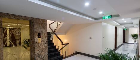 A modern hotel lobby with a marble reception desk, a staircase, and a hallway leading to the exit.
