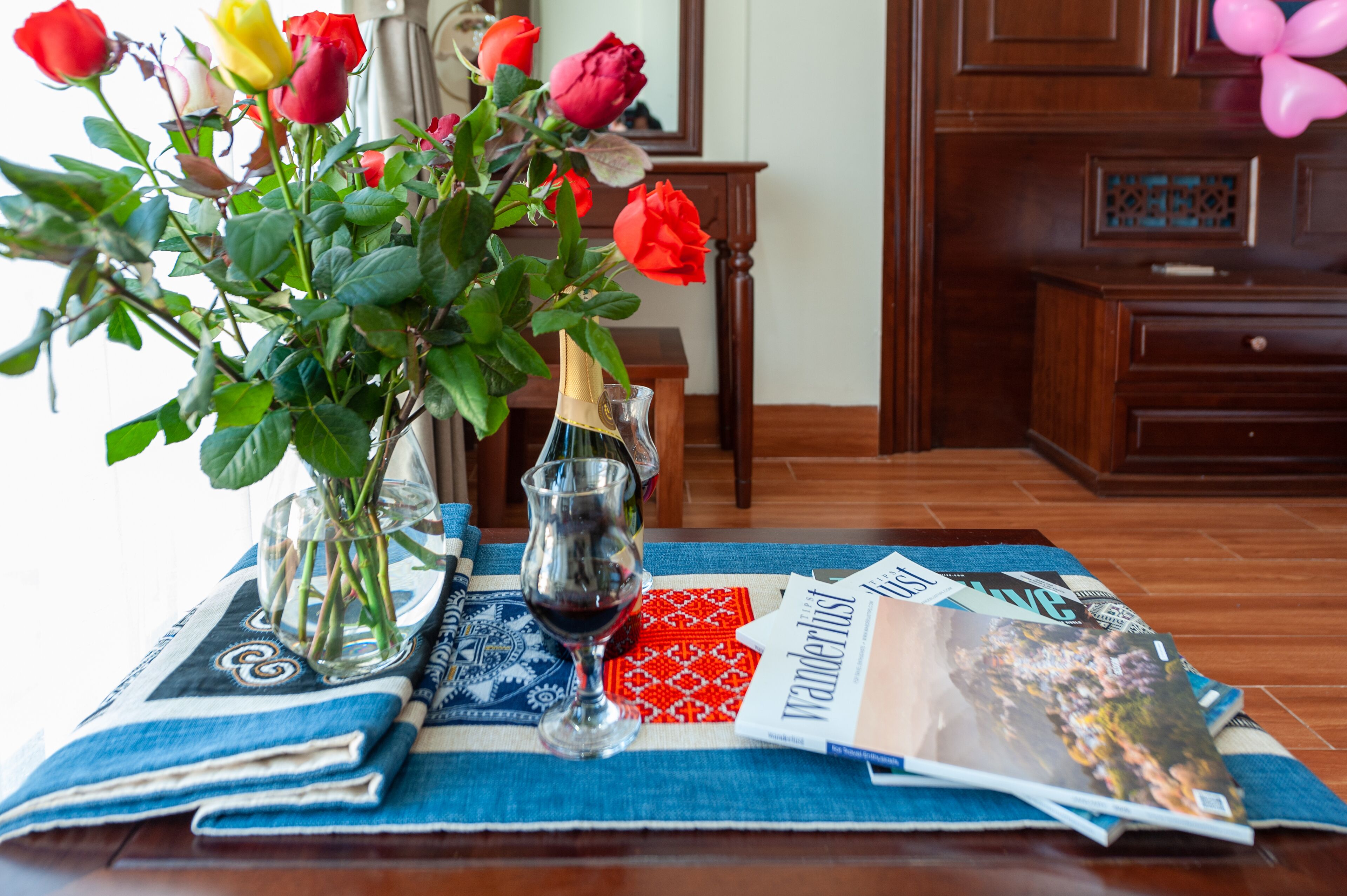 A table with a blue and white patterned tablecloth, a vase with roses, a wine glass, and a magazine titled 'Wanderlust'.