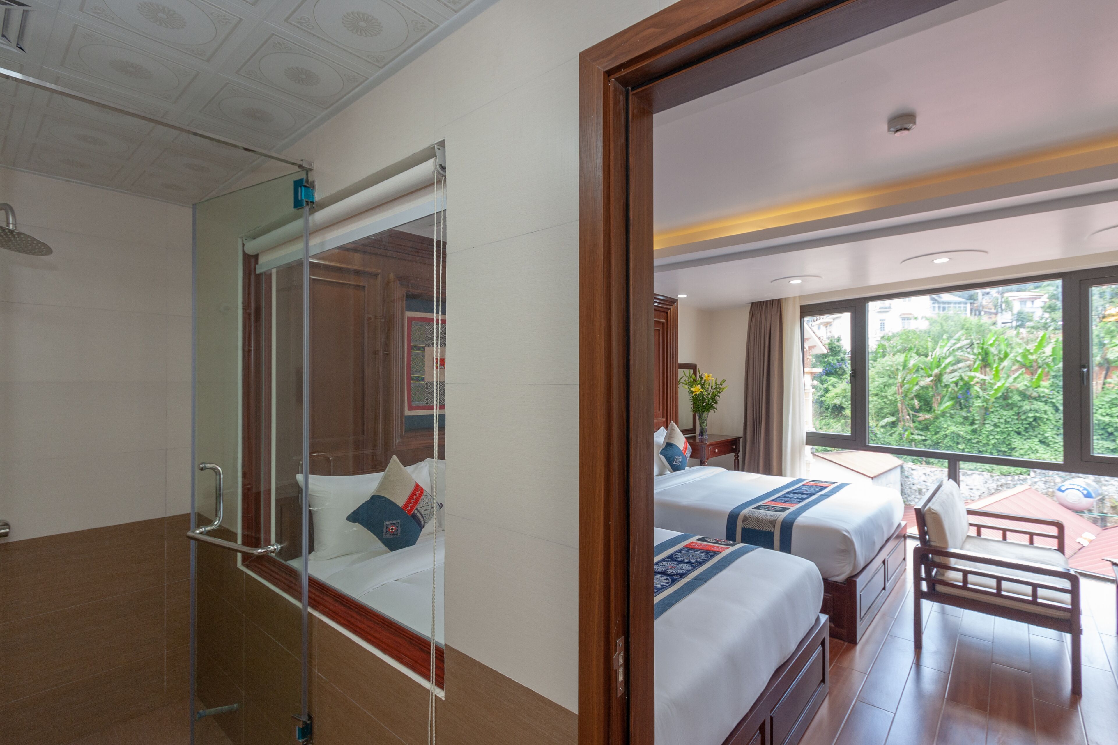 A hotel room with a shower, two beds, a small table, and a view of greenery outside.