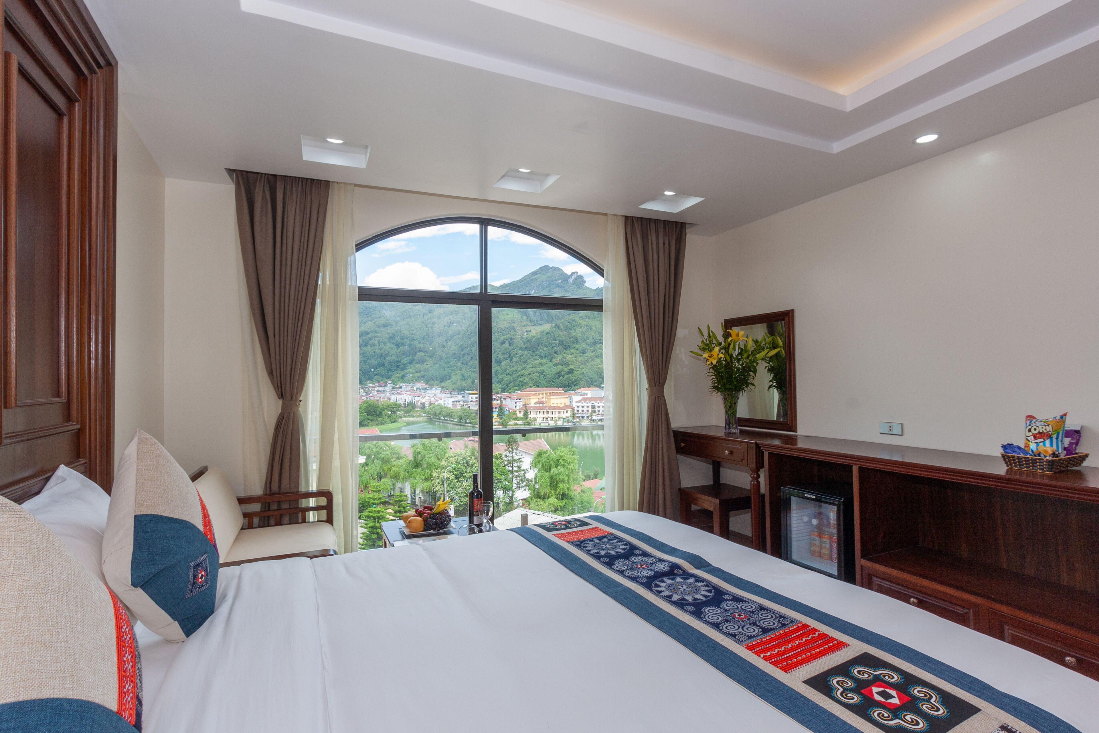A hotel room with a large bed, a desk, a chair, a small refrigerator, and a view of a mountain and city through a large window.