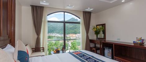 A hotel room with a large bed, a desk, a chair, a small refrigerator, and a view of a mountain and city through a large window.