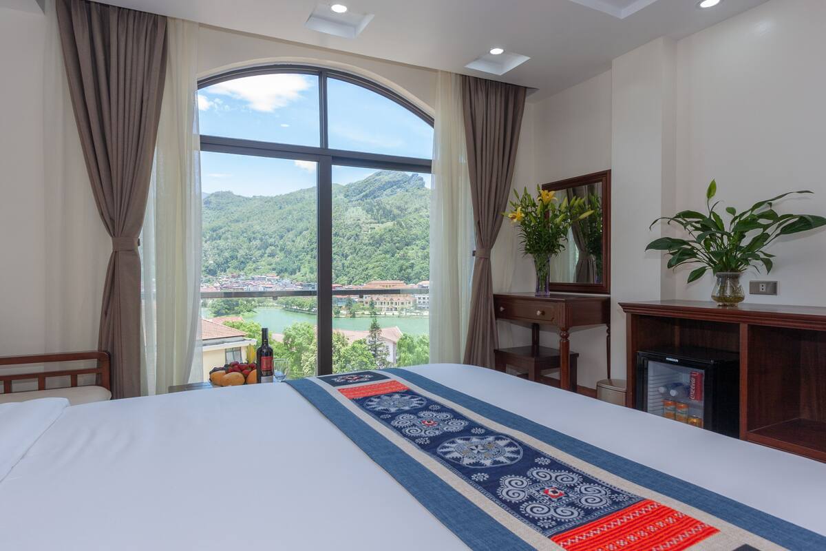 Quarto de hotel com uma grande janela que oferece vista para uma paisagem montanhosa e um rio. O quarto possui uma cama com um cobreje decorativo, uma mesinha com frutas e uma garrafa, e uma planta sobre uma mesa lateral.