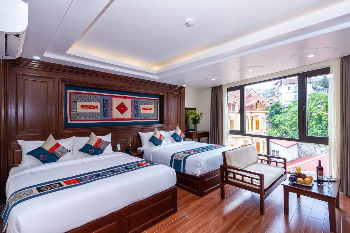 Quarto de hotel com duas camas, cabeceira de madeira, uma janela grande com vista para edifícios e uma pequena mesa com fruta e uma garrafa.