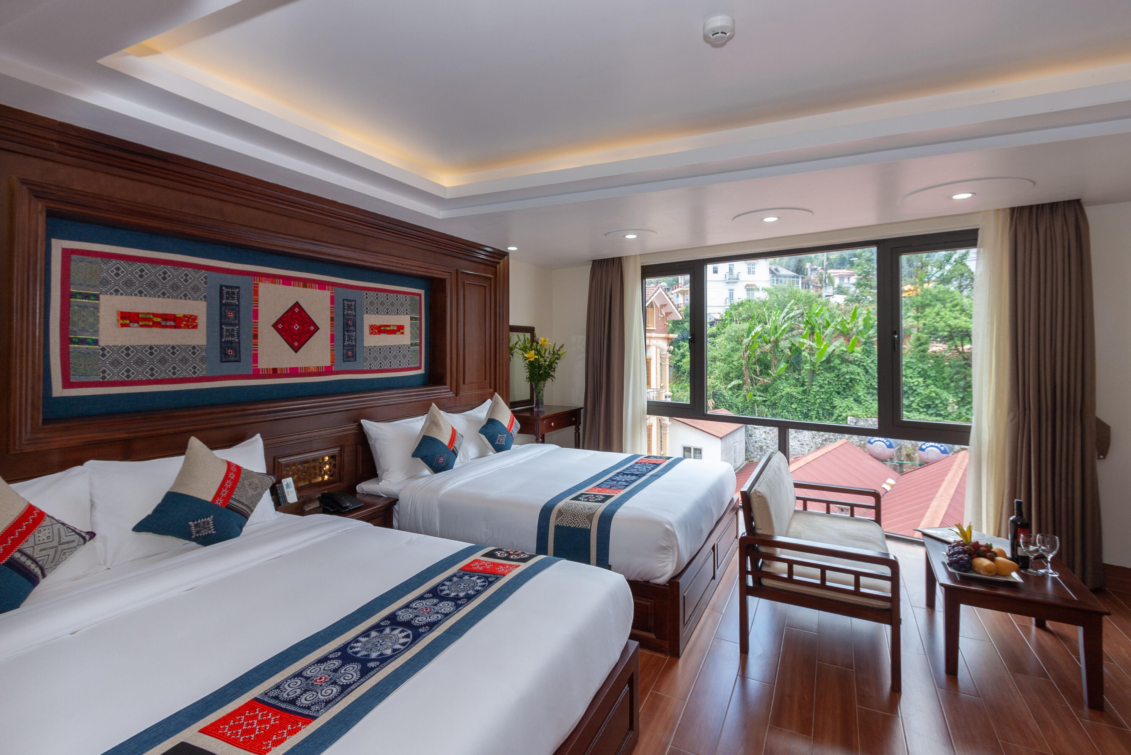 A hotel room with two beds, a wooden headboard, a dining table with fruit, and a view of greenery outside.