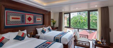 A hotel room with two beds, a wooden headboard, a dining table with fruit, and a view of greenery outside.