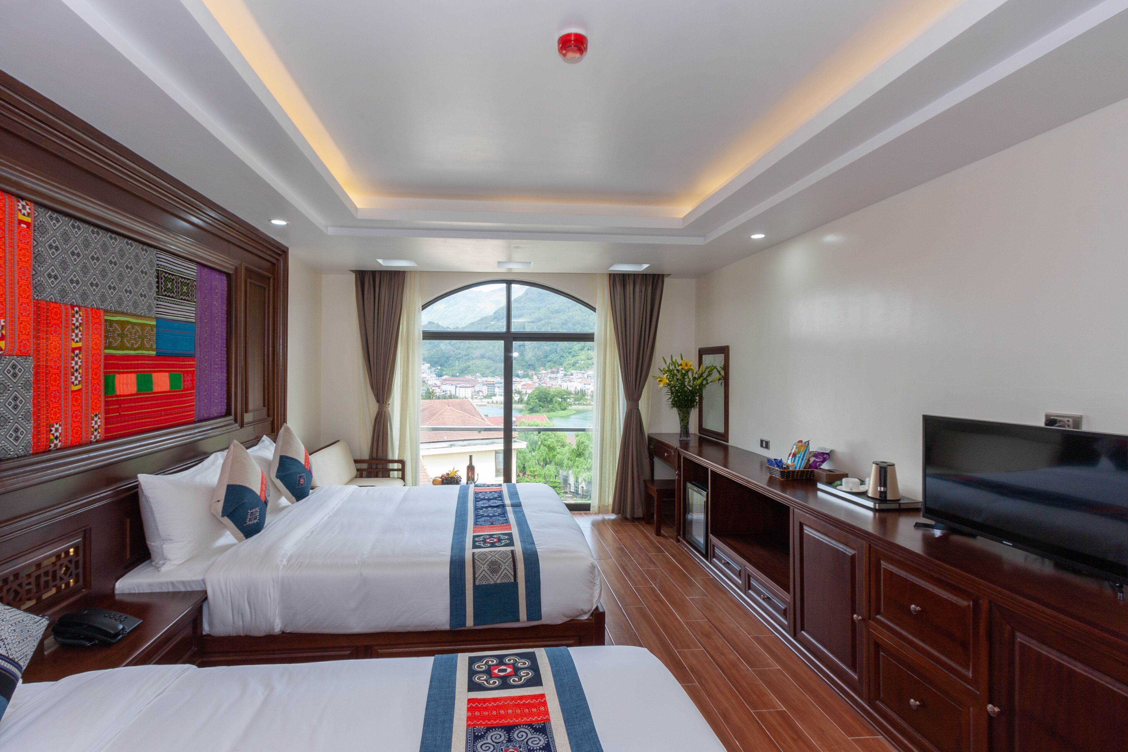 A hotel room with two beds, a TV, a desk, and a large window with a view of the city.