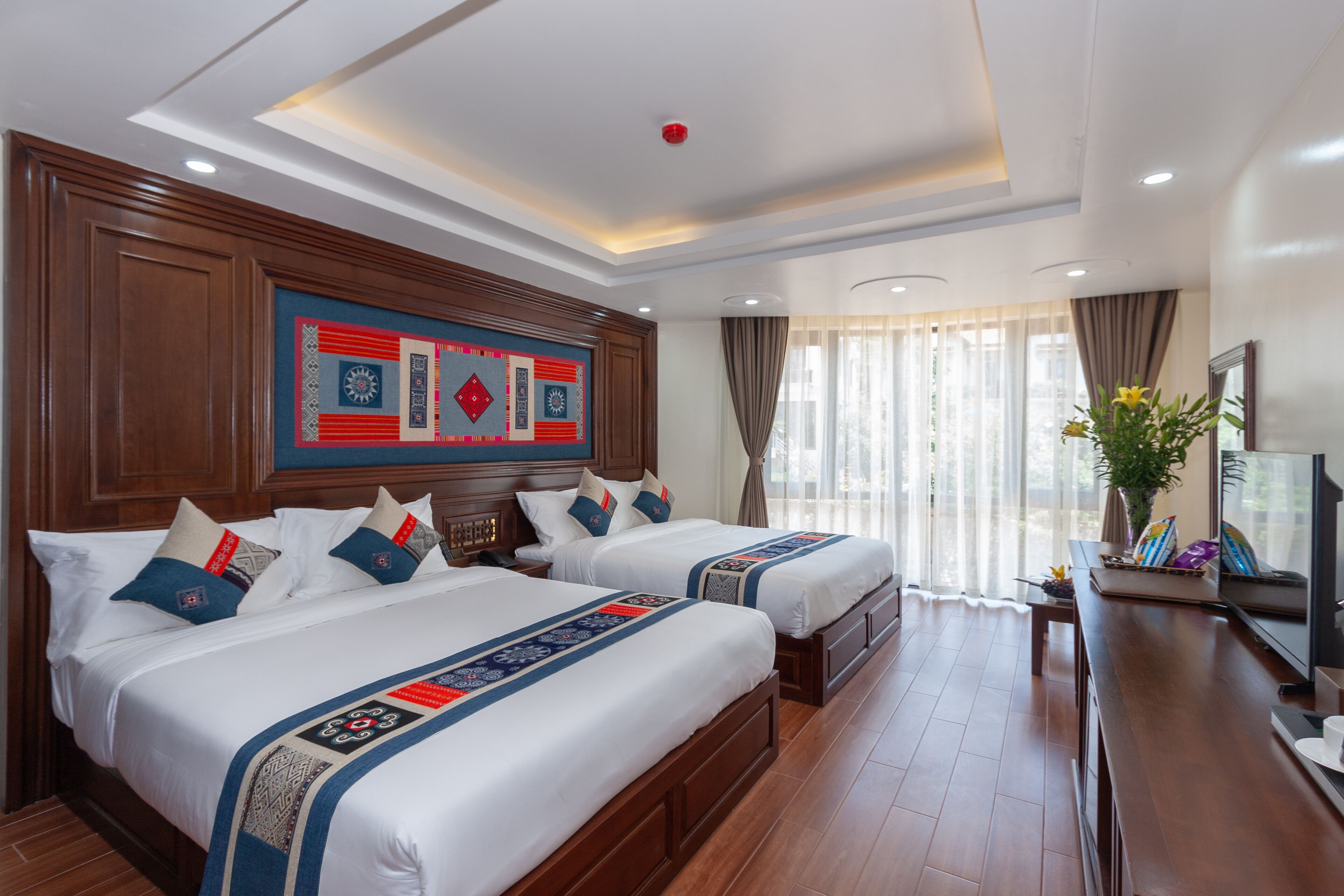 Une chambre d’hôtel avec deux lits, des têtes de lit en bois, un grand élément décoratif mural, un téléviseur à écran plat, un bureau avec un ordinateur portable et une plante en pot.