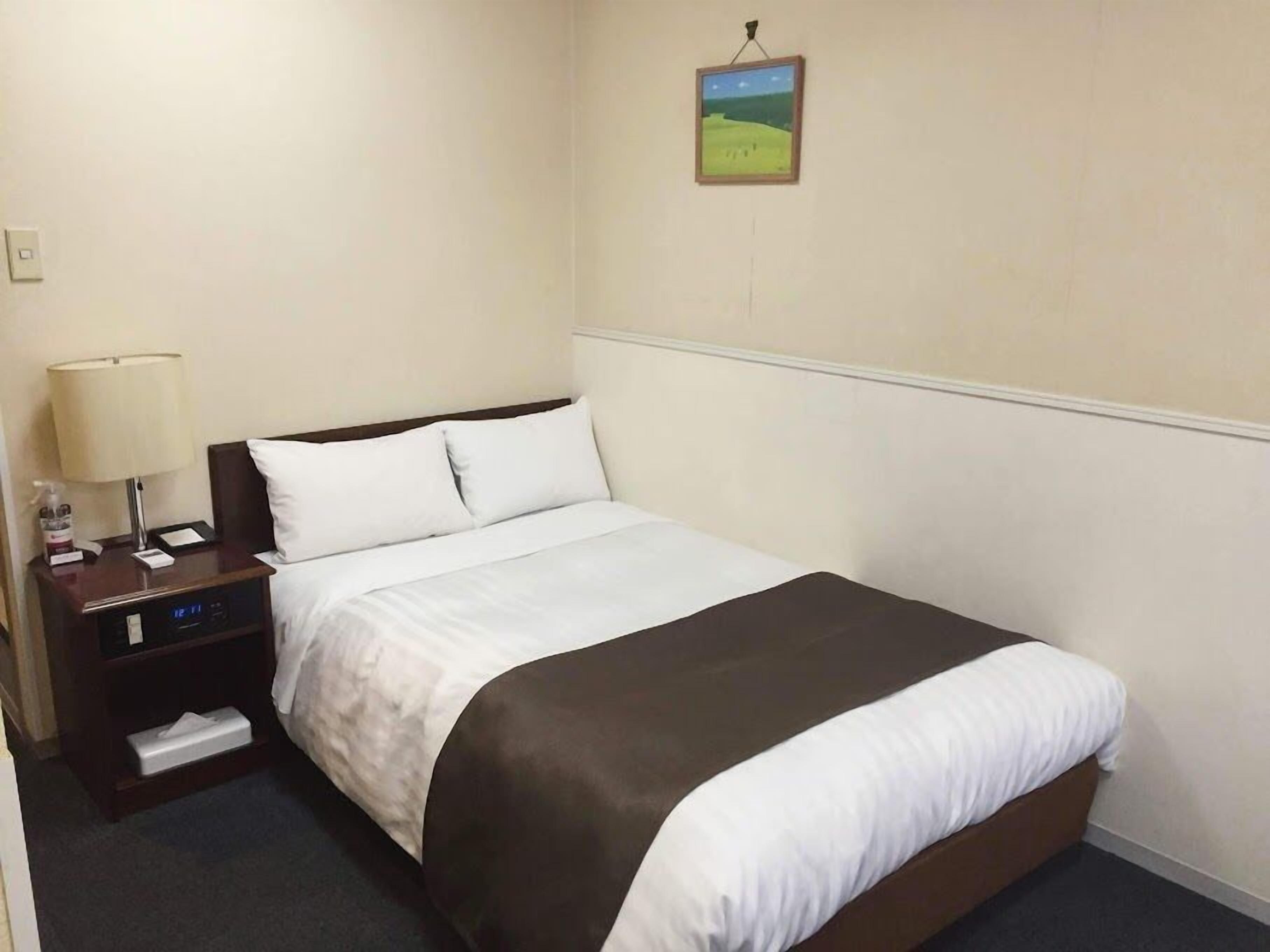 A hotel room with a bed, a nightstand with a lamp, a digital clock, and a small painting on the wall.