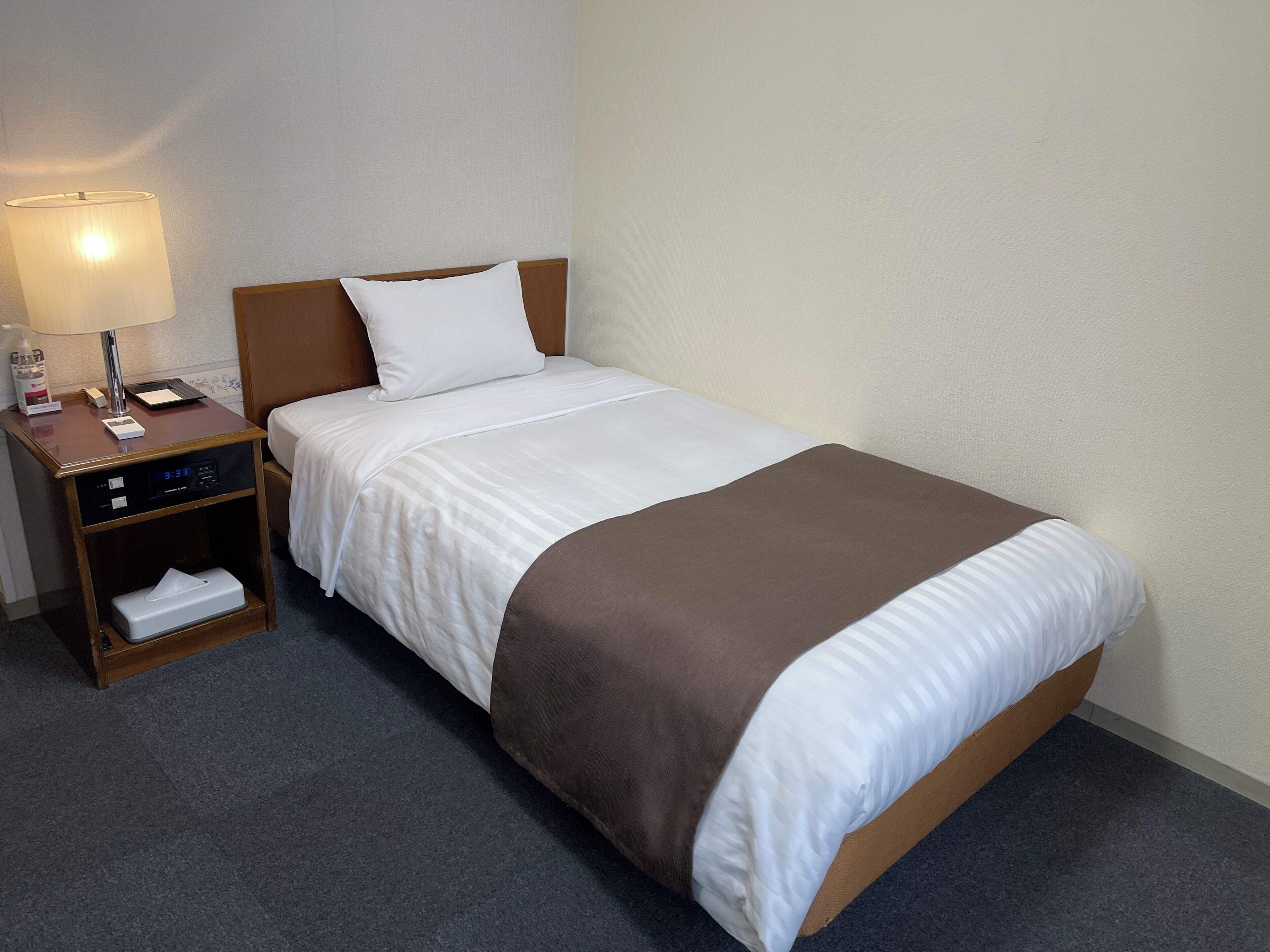 A neatly made bed with a brown blanket, a white pillow, and a wooden headboard. A small bedside table with a lamp and a digital clock is visible.